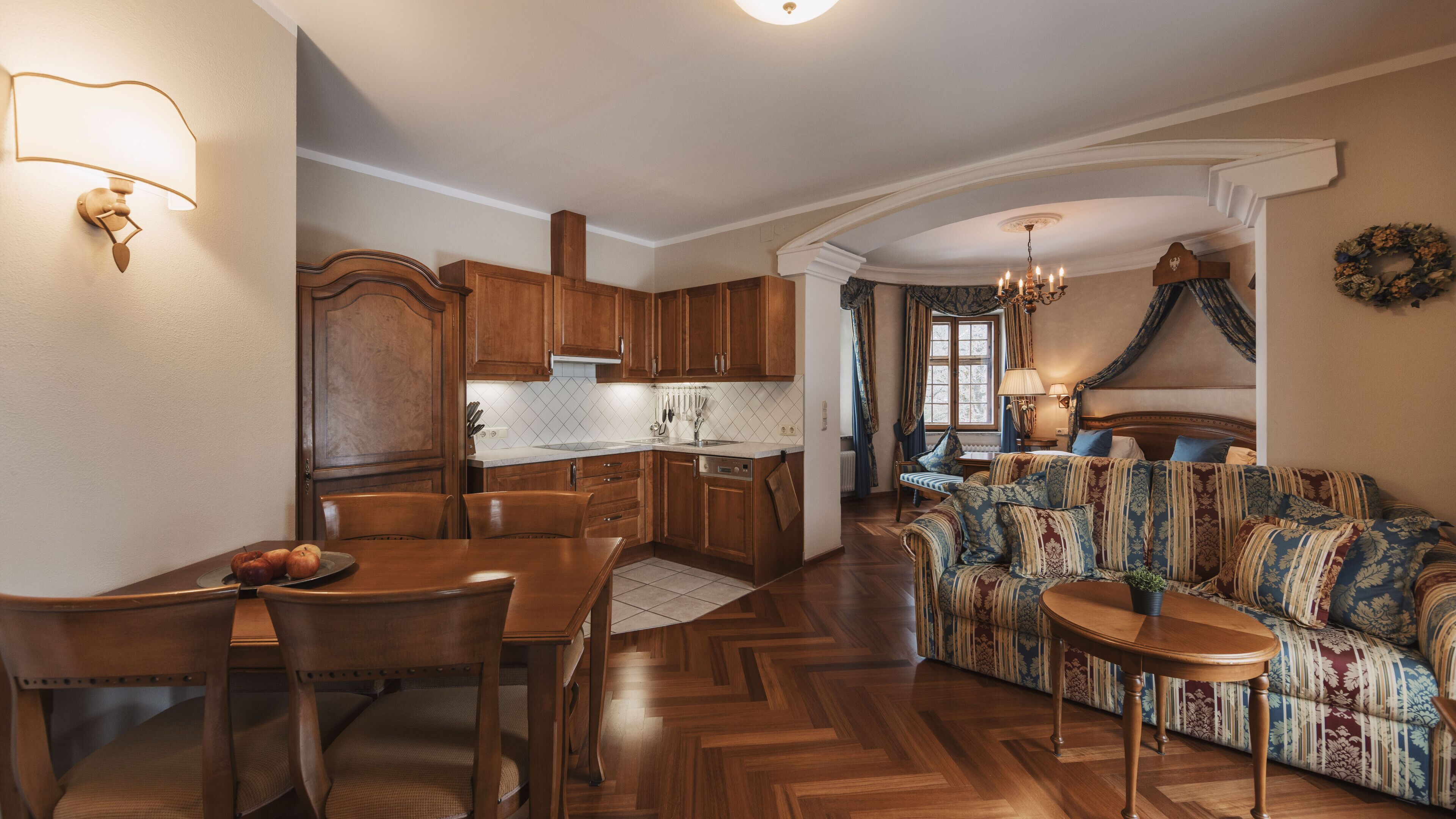 Un salón acogedor con un sofá estampado, una mesa de comedor de madera y sillas. Se ve una cocina con armarios de madera y una ventana al fondo.