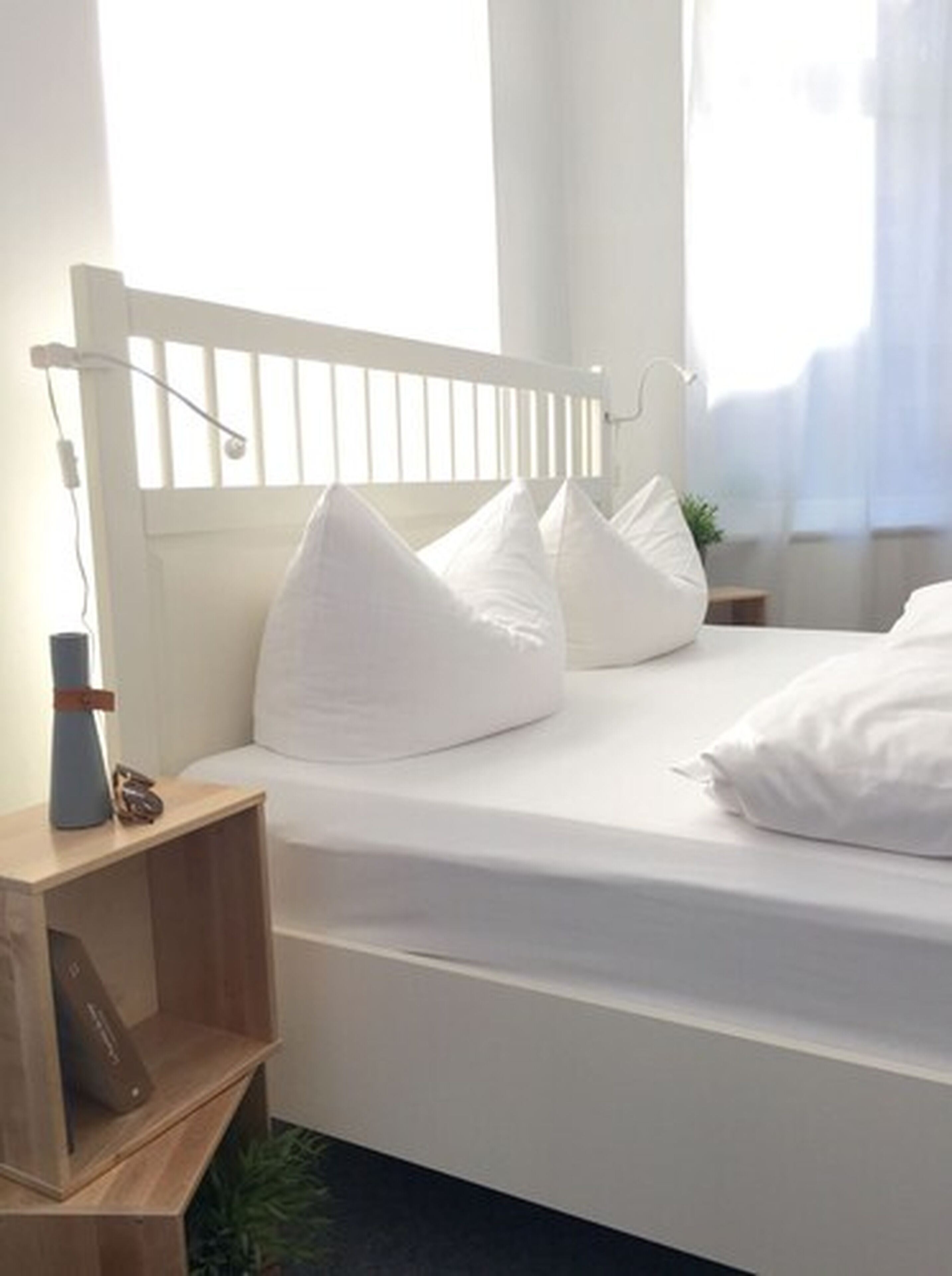 Una cama blanca con cabecera de madera, una mesita de noche con una lámpara y una planta pequeña.