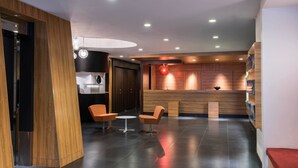 Un espace d’accueil moderne avec un comptoir de réception en bois, deux fauteuils orange, une petite table et un luminaire sphérique rouge.