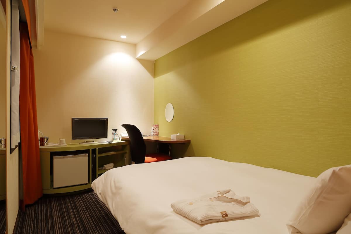 A hotel room with a bed, a desk with a computer, a chair, a small refrigerator, and a window with curtains.