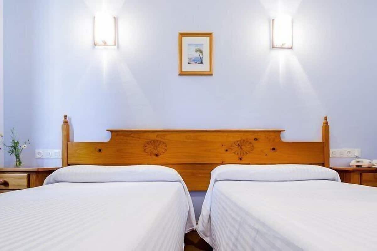 Quarto de hotel com duas camas de solteiro, cabeceira de madeira, duas mesas de cabeceira, um criado-mudo, um quadro emoldurado na parede e duas luzes fixadas na parede.