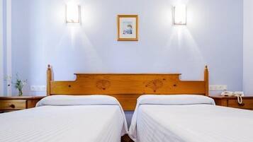 Una habitación de hotel con dos camas individuales, cabecera de madera, dos mesitas de noche y dos lámparas de pared. También se observa un cuadro enmarcado en la pared.