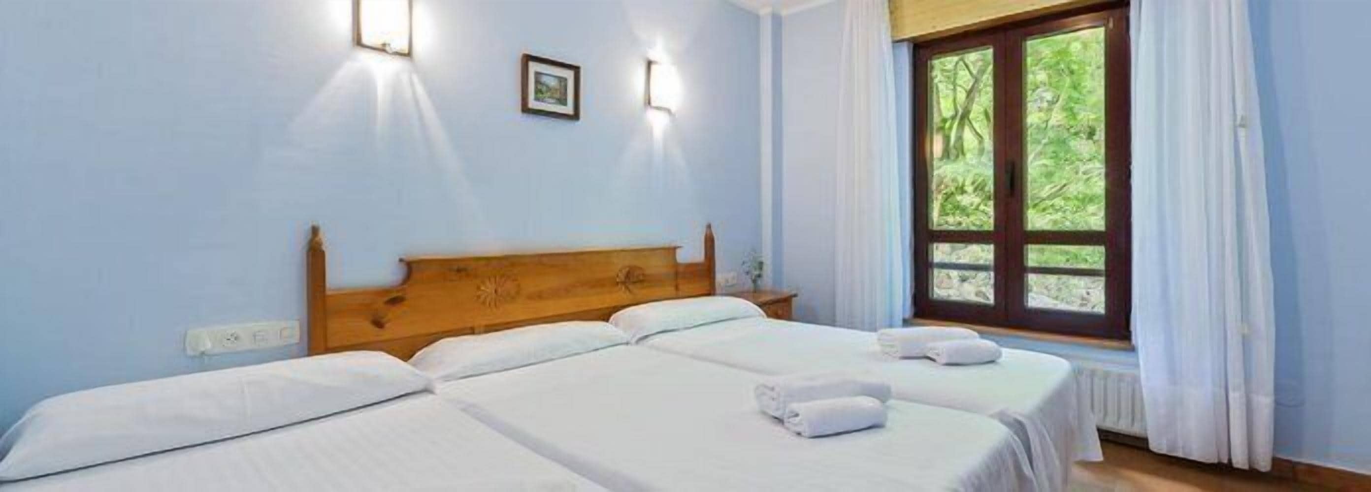 Une chambre d’hôtel avec deux lits simples, une tête de lit en bois, une fenêtre avec des rideaux et un luminaire fixé au mur.