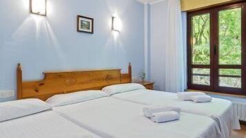 Habitación de hotel con dos camas individuales, cabecero de madera, ventana con cortinas e iluminación fijada a la pared.