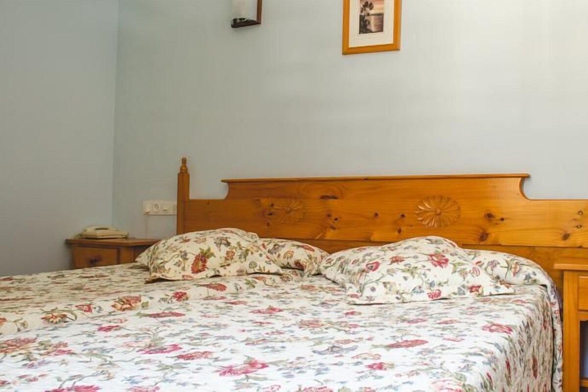 Una cama individual con ropa de cama floral, cabecera de madera, mesita de noche con lámpara y un aparador con teléfono.