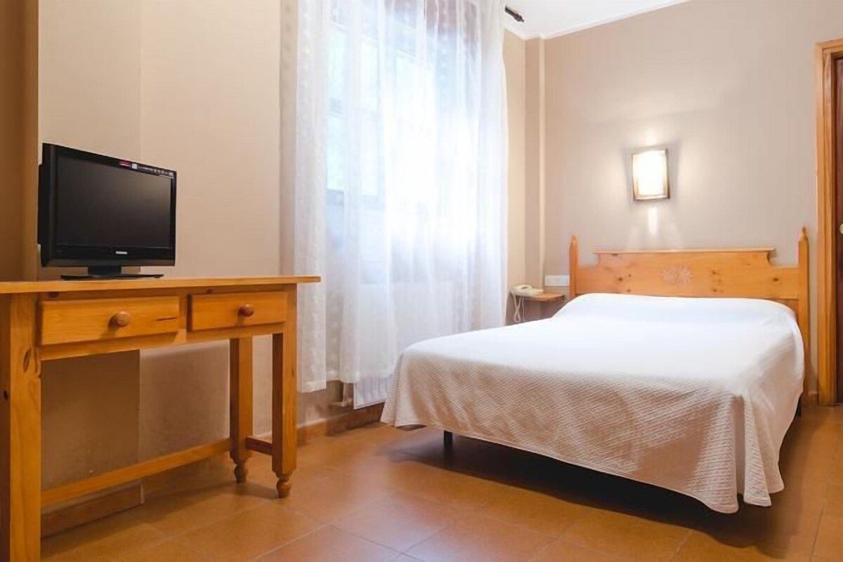 Una habitación de hotel con cama, un escritorio de madera con televisión, una mesita de noche y una puerta.