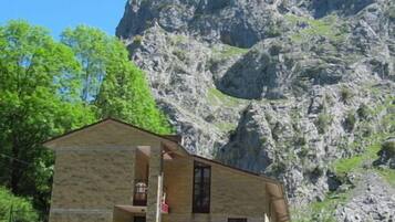 A stone building with a balcony, surrounded by greenery and a rocky mountain backdrop.