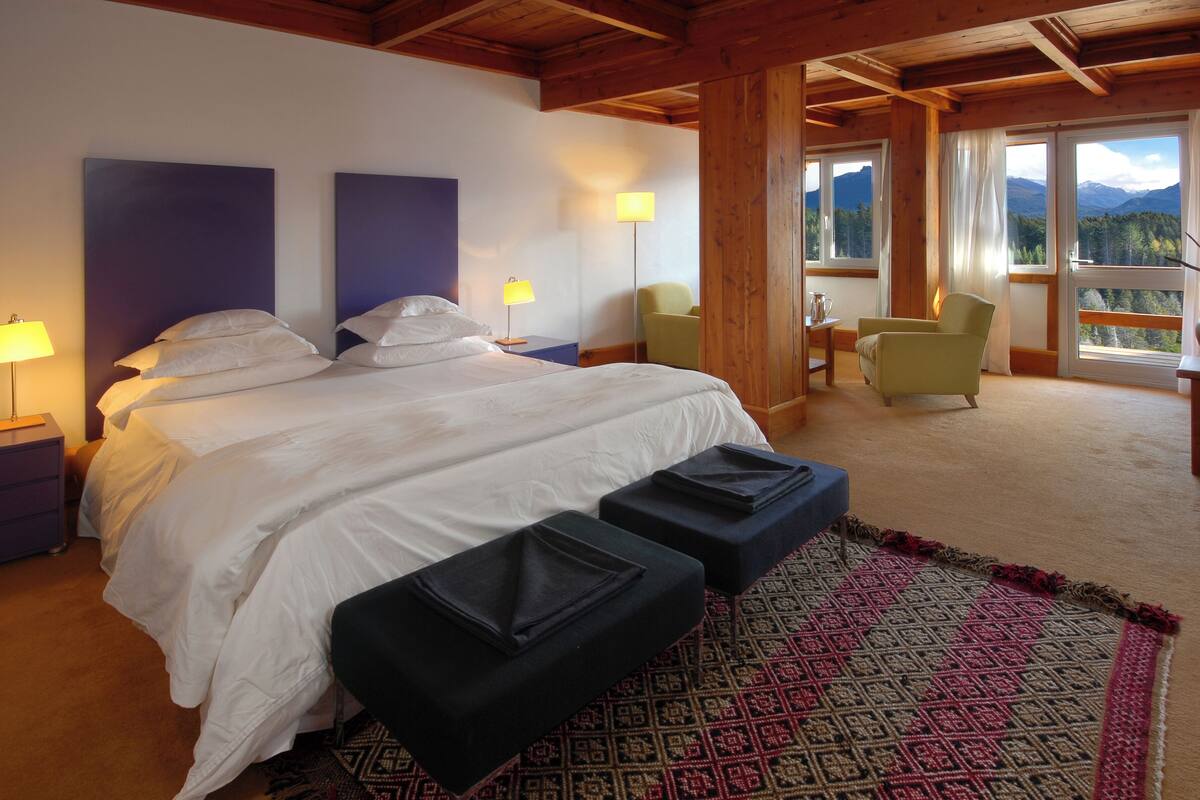 A bedroom with a large bed, two bedside lamps, a chair, a table, and a view of mountains through the window.
