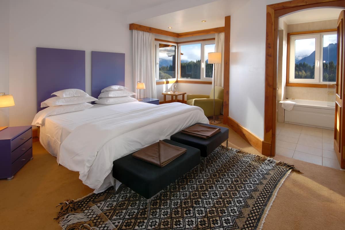 A hotel room with a large bed, a desk, a chair, a bathroom with a bathtub, and a view of mountains through the windows.