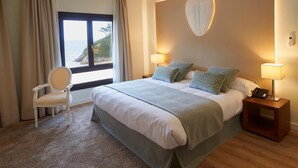 Quarto de hotel com uma cama grande, duas luminárias de cabeceira, uma cadeira e vista para o mar através de uma janela ampla.