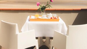 Mesa com toalha branca, duas cadeiras brancas, um vaso com flores cor-de-rosa e um tabuleiro com comida e bebidas. A mesa está colocada num deck de madeira com vista para a praia e para falésias.