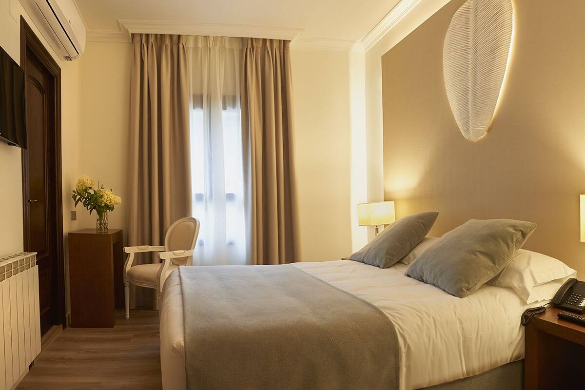 Habitación de hotel con cama, lámparas de noche, escritorio con teléfono, silla, radiador y televisor en la pared.
