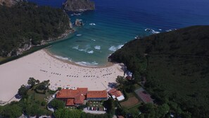 An aerial view of a beach with a large building complex, including a hotel with multiple floors and a tennis court.