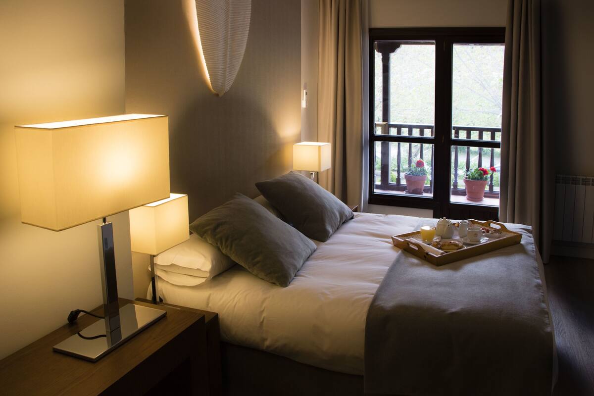 Una cama bien hecha con almohadas, dos lámparas de noche, una bandeja con alimentos para el desayuno y una ventana con plantas en macetas.