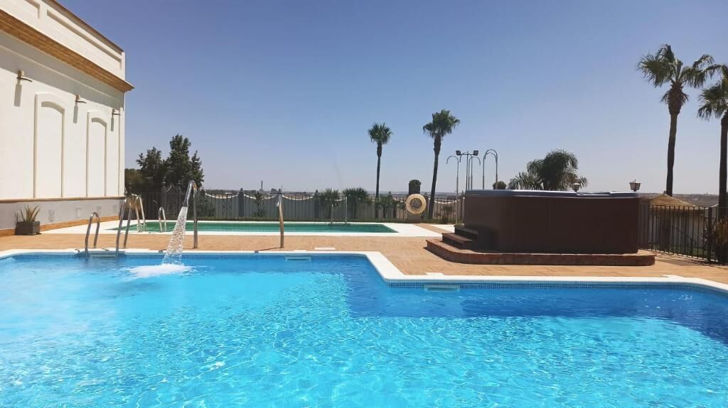 Una piscina con agua cristalina azul, rodeada por una valla, palmeras y un edificio con fachada blanca.