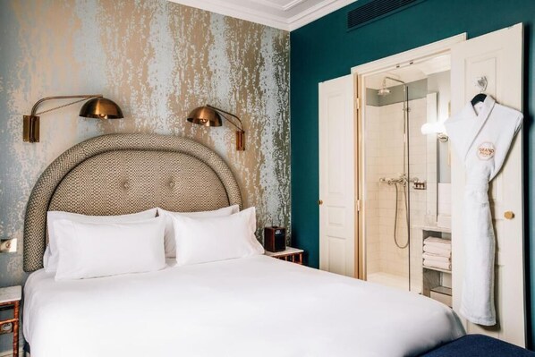 A hotel room with a large bed, a headboard with a geometric pattern, two wall-mounted lamps, a wooden nightstand, a robe hanging on the door, and a shower area visible through an open door.