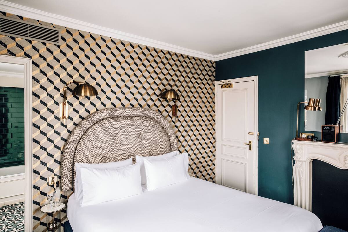 A bedroom with a patterned wallpaper, a large bed with white bedding, two bedside lamps, and a white door.