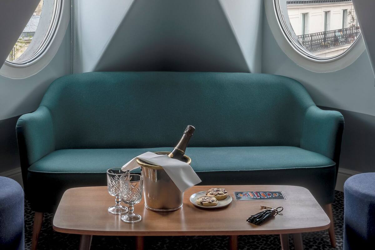 A cozy room with a teal sofa, a small table with a bottle, glasses, and a plate of cookies. There are two windows with views of buildings.