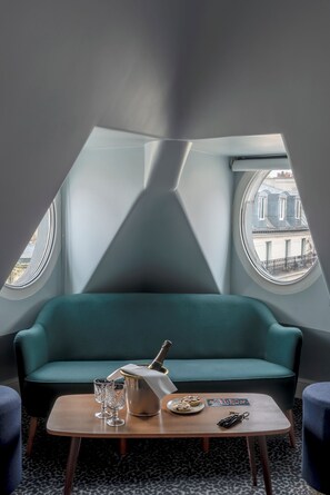 Un cuarto acogedor con un sofá color turquesa, una mesita con una botella, vasos y un plato de galletas. Hay dos ventanas con vistas a edificios.