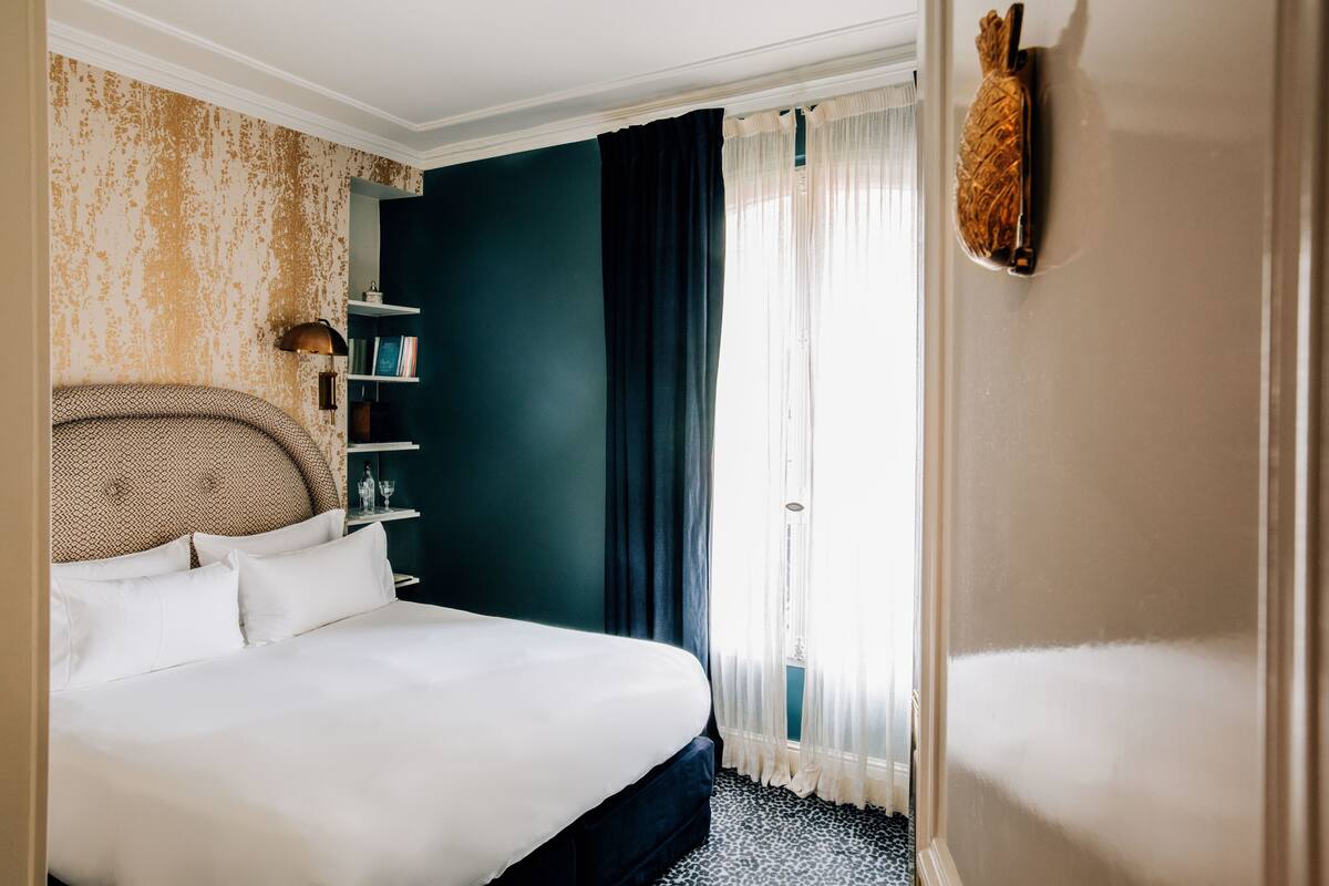A neatly made bed with white linens, a dark blue wall, and a bookshelf with books and decorative items.