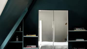 A modern hotel room with a mirrored wardrobe, a bed, a small table with books and glasses, and a blue ottoman.