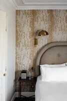 Ett hotellrum med tapet med ett mönster, en säng med vita sängkläder, ett nattduksbord med en radio, ett glas och en lampa i mässing.