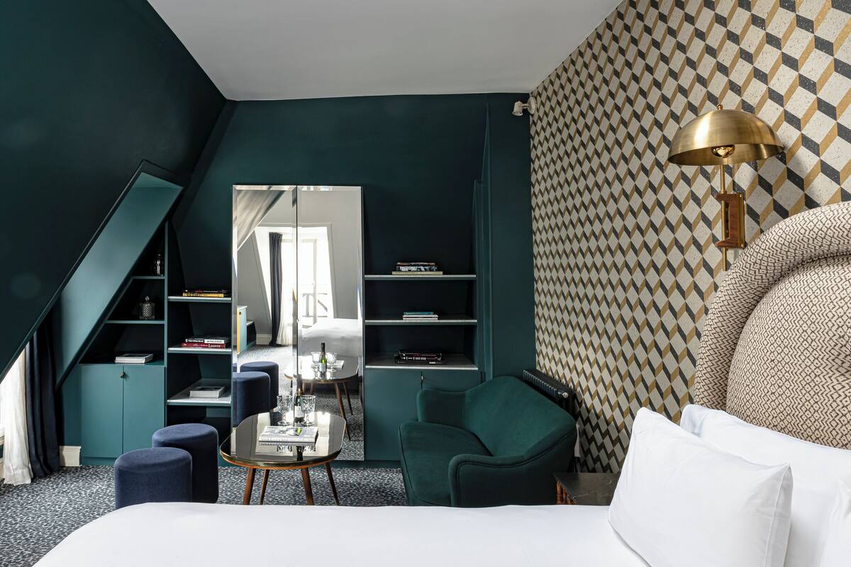 A modern hotel room with a sloped ceiling, a large bed, a desk with a chair, a green armchair, and a bookshelf.