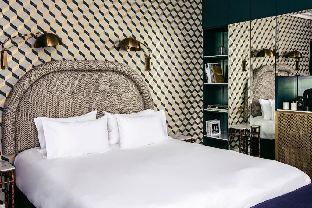 Een keurig opgemaakt bed met witte lakens, een hoofdbord met een geometrisch patroon en twee bedlampjes.