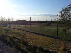 A tennis court surrounded by a chain-link fence, with a building in the background and sparse vegetation in the foreground. - Borgo Beccanella (Asciano)