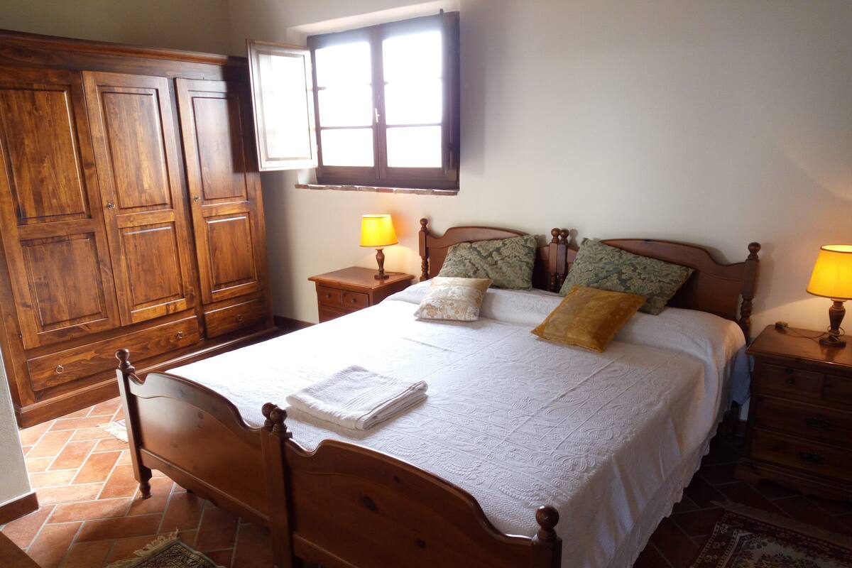 A bedroom with a wooden bed, two bedside tables with lamps, a window with curtains, and a wooden wardrobe.