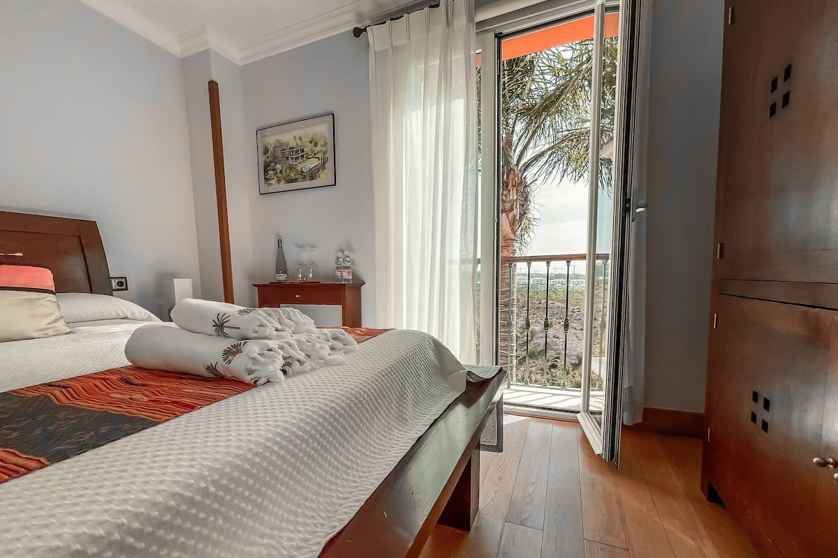 Een hotelkamer met een bed, nachtkastje, schilderij en een balkon met uitzicht.