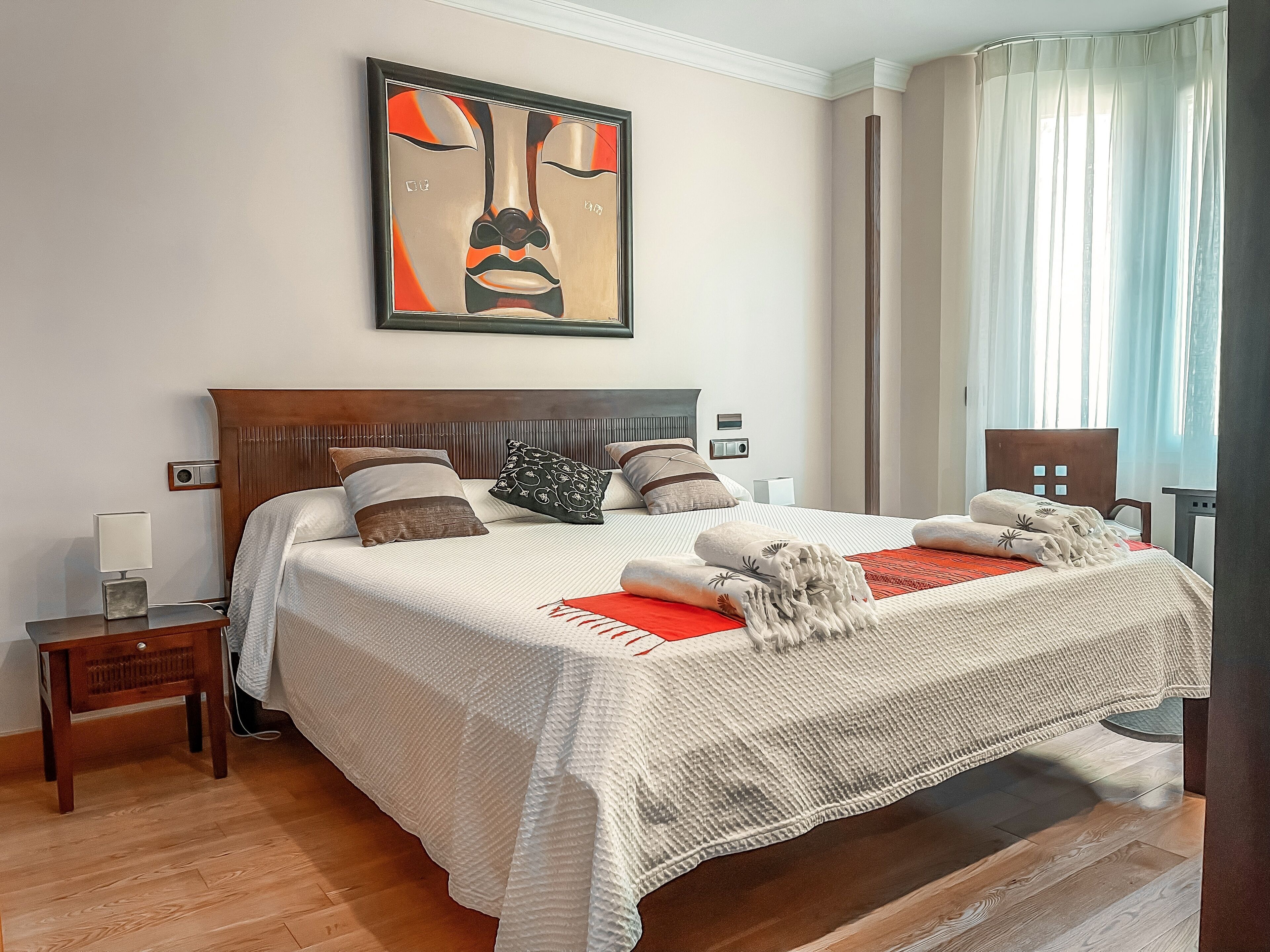 Un lit bien fait, recouvert d’une couette blanche, agrémenté de plusieurs oreillers et d’un plaid rouge. Une table de chevet avec une lampe et une radio se trouve à côté du lit. La pièce est dotée d’un parquet et d’une grande fenêtre ornée de rideaux légers.