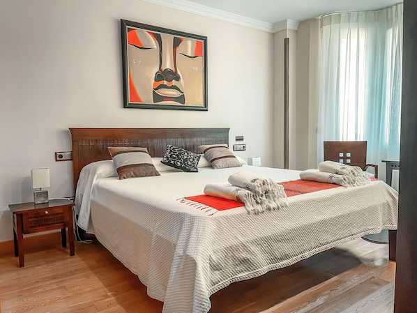 A neatly made bed with a white duvet, multiple pillows, and a red throw blanket. There is a bedside table with a lamp and a radio. The room has wooden flooring and a large window with sheer curtains. - The Marbella Heights Boutique Hotel (Marbella)