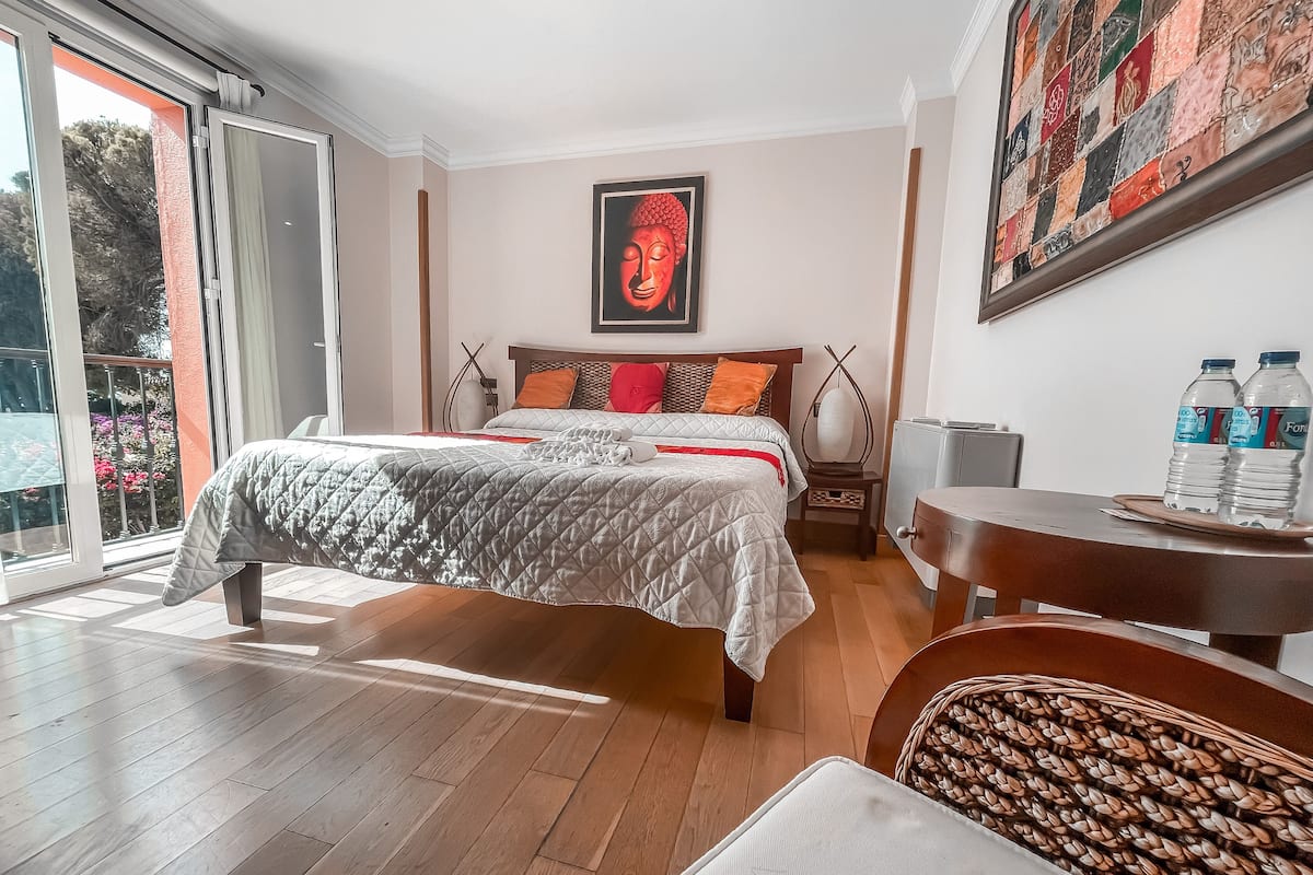 Een slaapkamer met een groot bed, een houten vloer, een raam, een schilderij en een tafel met waterflessen.