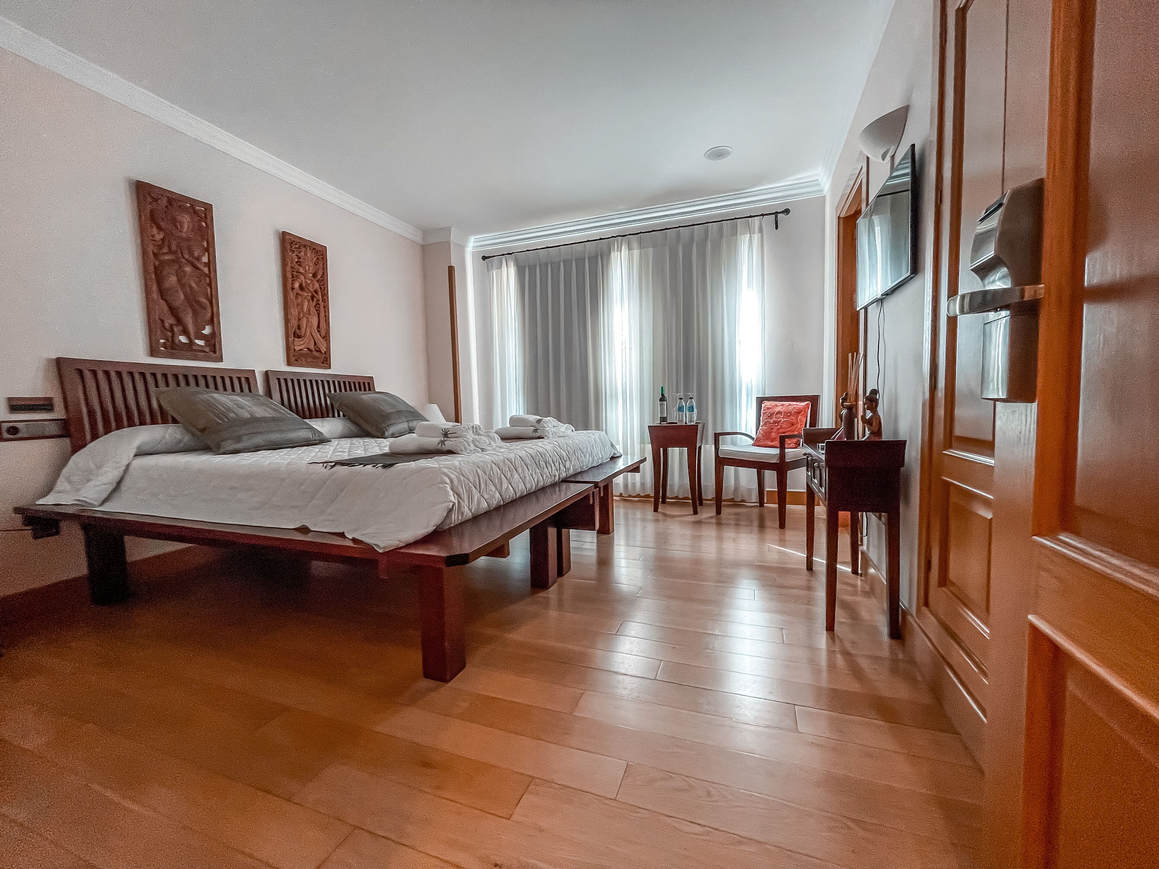 Habitación de hotel con cama de madera, una mesita con una silla, televisor montado en la pared y un ventanal con cortinas.