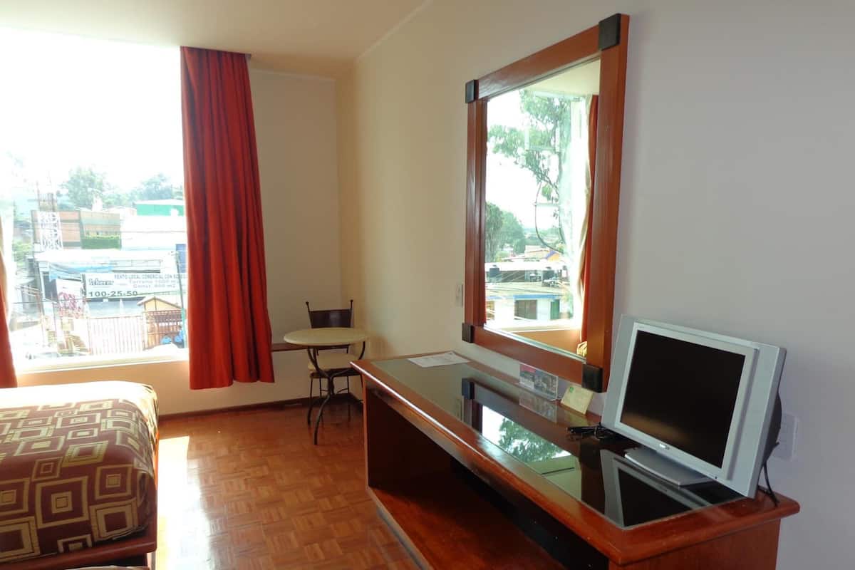 Une chambre d’hôtel avec un lit, un bureau équipé d’un ordinateur, une chaise et une fenêtre donnant sur des immeubles.