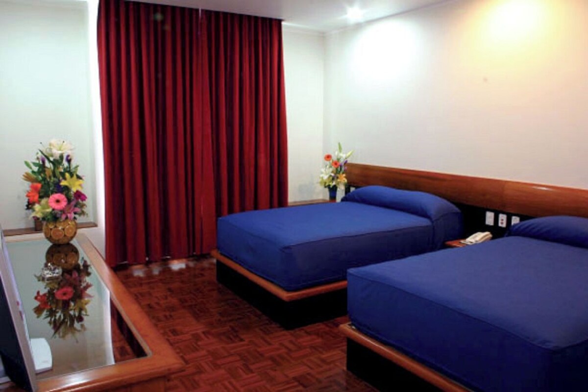 A hotel room with two beds, a wooden headboard, a red curtain, a small table with a vase of flowers, and a wooden floor.