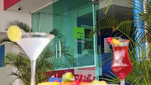 Entrada de hotel com porta de vidro, uma placa com a inscrição "GS Hotels" e uma mesa com uma taça de martini e frutas coloridas.