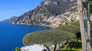 Un pueblo costero con una montaña destacada al fondo, un mar azul transparente y un jardín cuidado con una mesa y sillas.