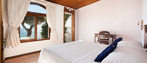 A bedroom with a large bed, a wooden ceiling, a window with curtains, and a view of trees.