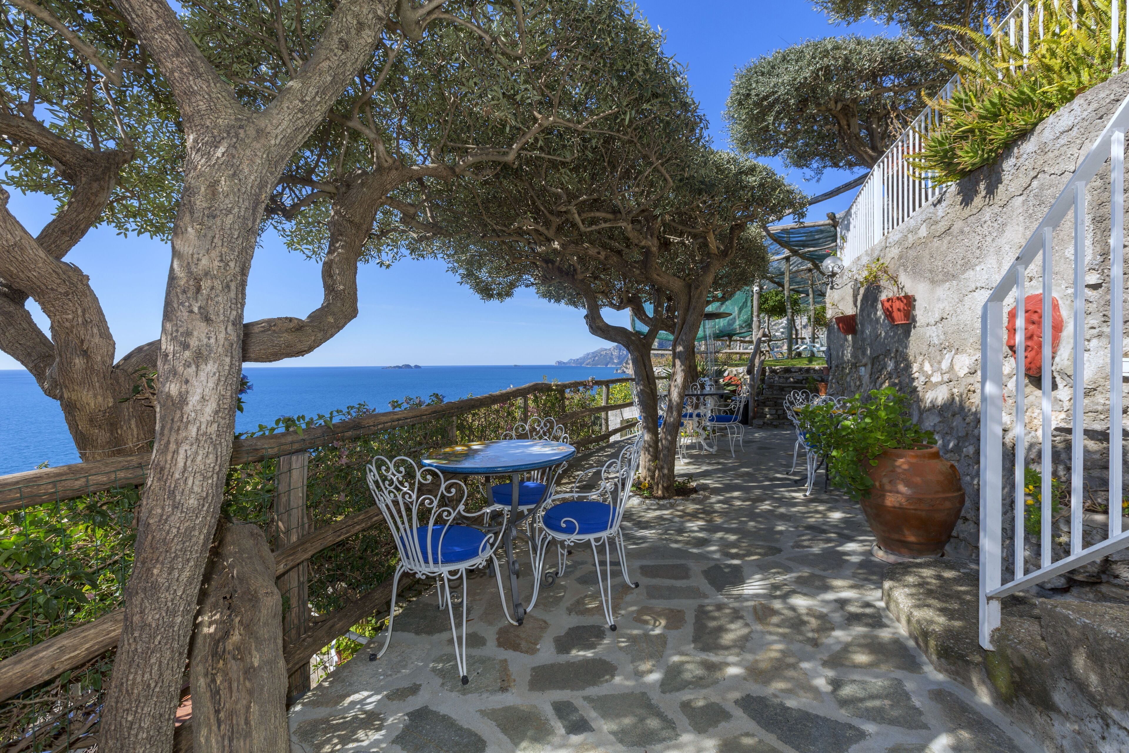 A stone-paved outdoor seating area with metal chairs and a table, overlooking a coastal view.