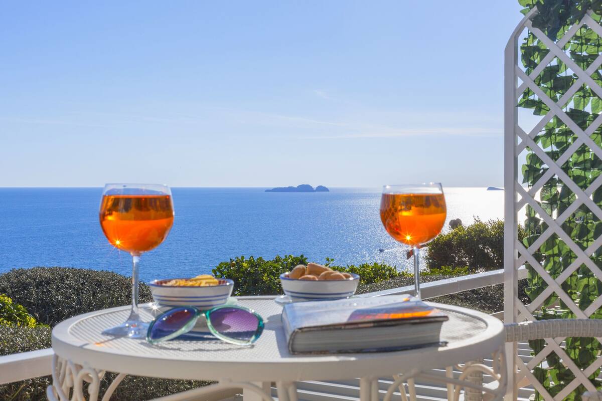 Una mesa con dos vasos de bebida de naranja, un plato de comida y un libro, frente a una vista panorámica del mar y una isla.