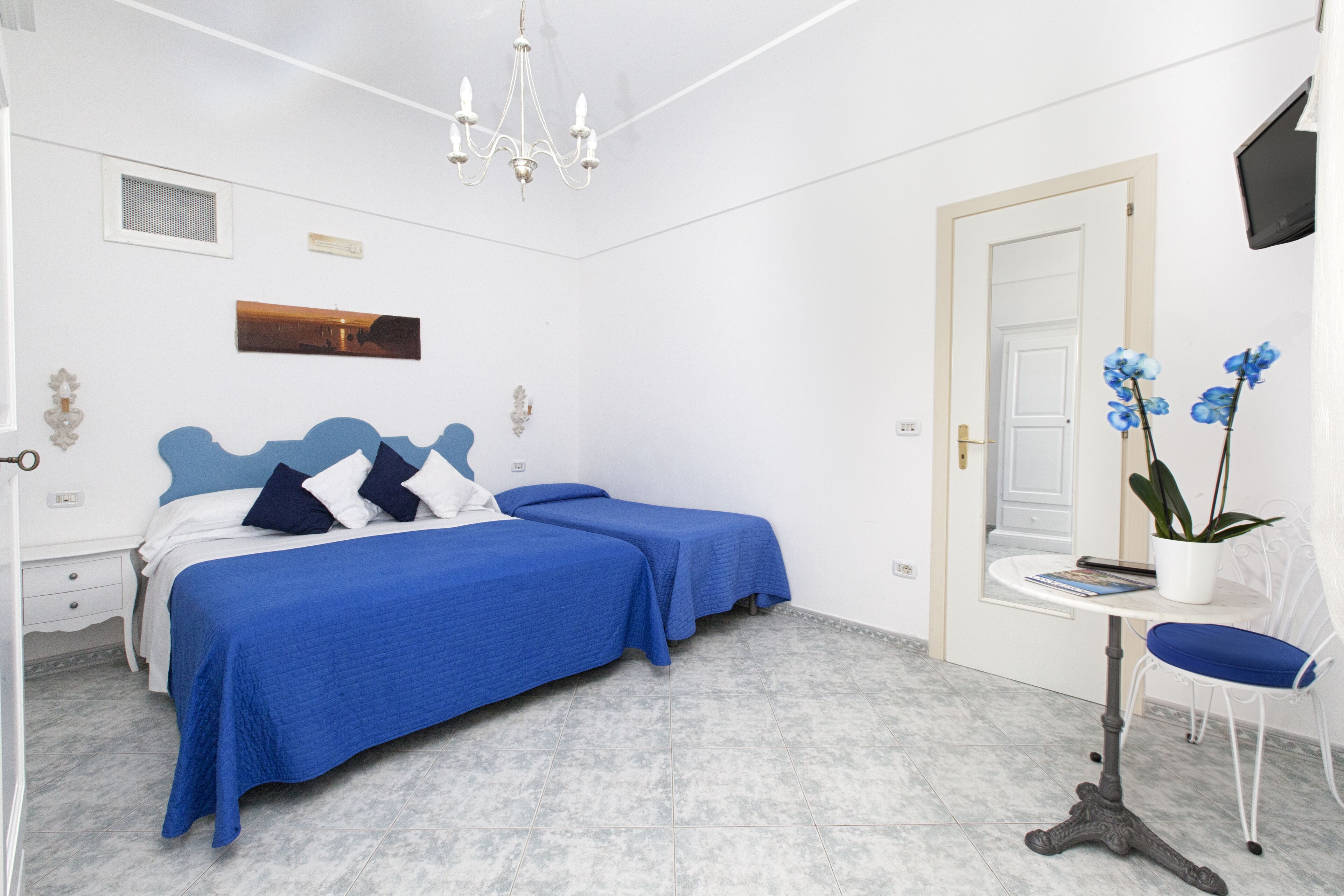 A bedroom with two beds, a small table, a chair, a potted plant, and a TV on the wall.
