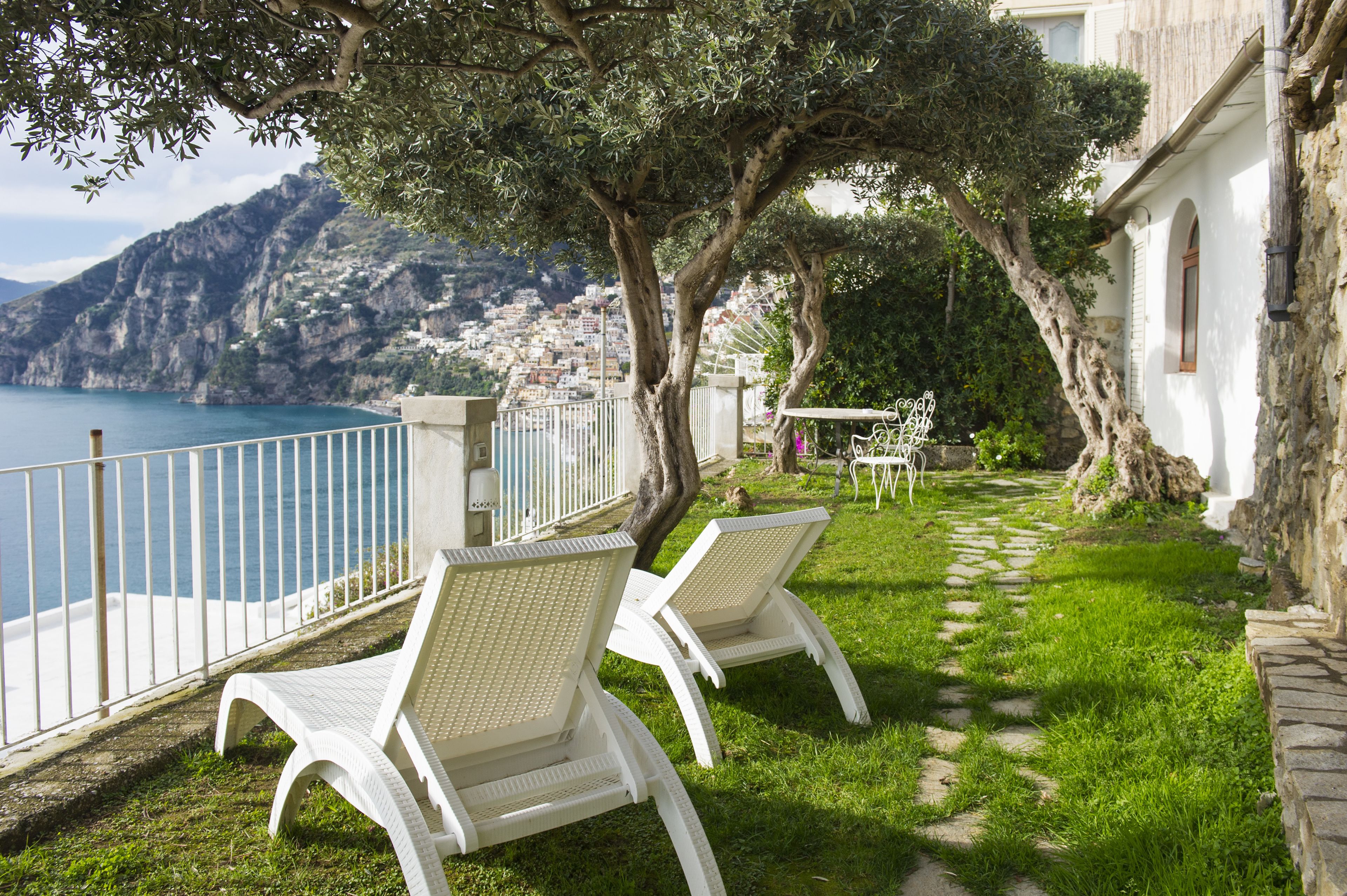 A terrace with white chairs, a stone pathway, and a view of the sea and mountains.