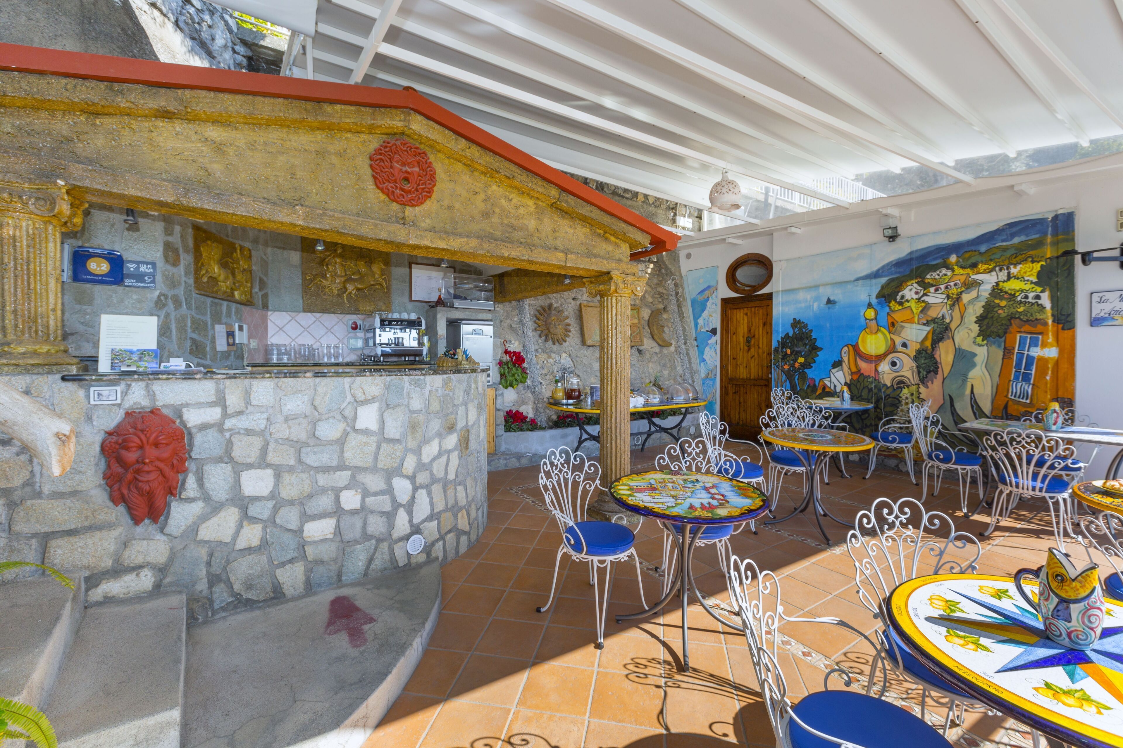 An outdoor café with stone walls, metal chairs, and tables with colorful tablecloths.