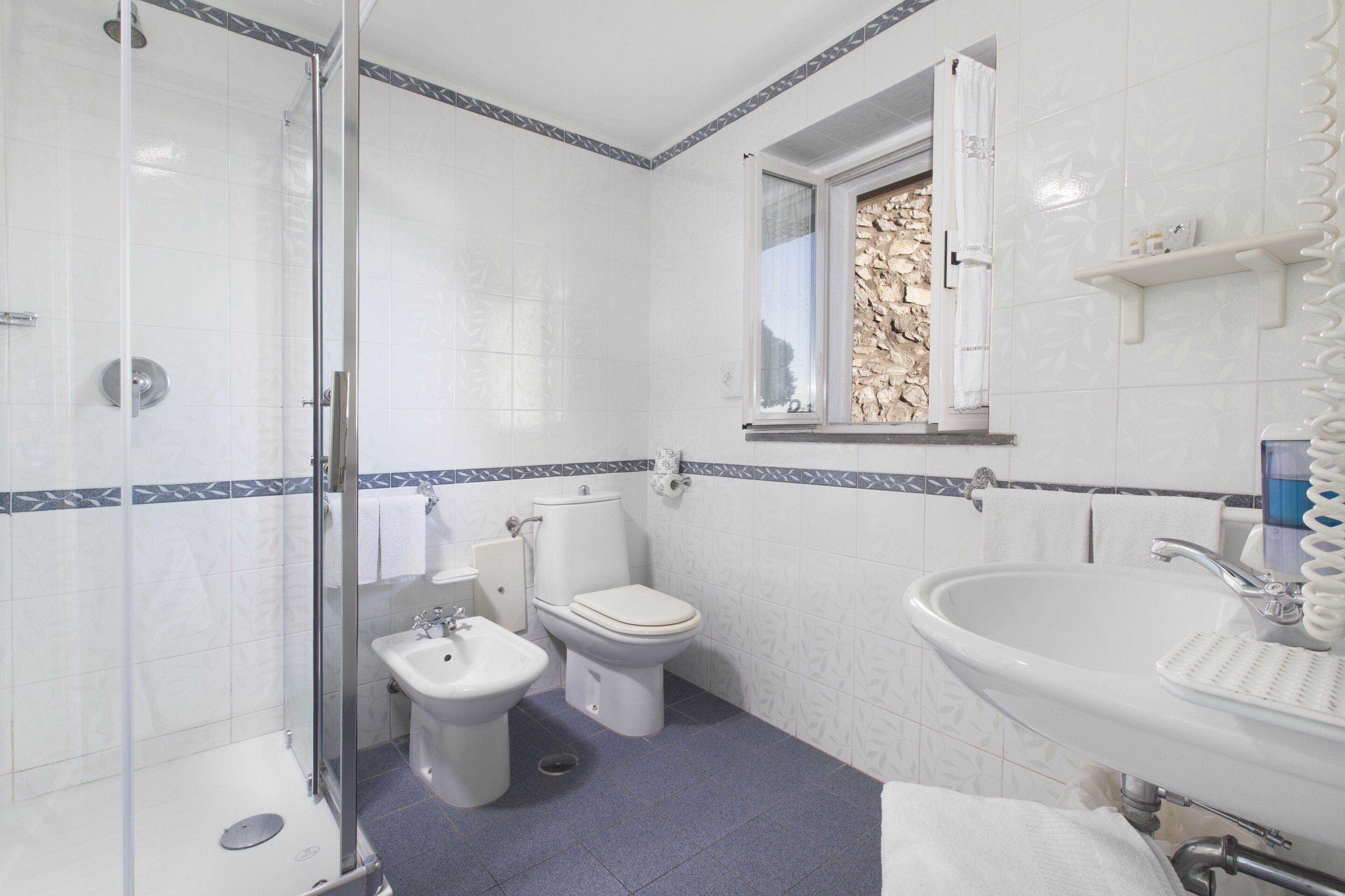 A bathroom with a glass shower enclosure, a bidet, a toilet, and a sink with a towel rack above it.