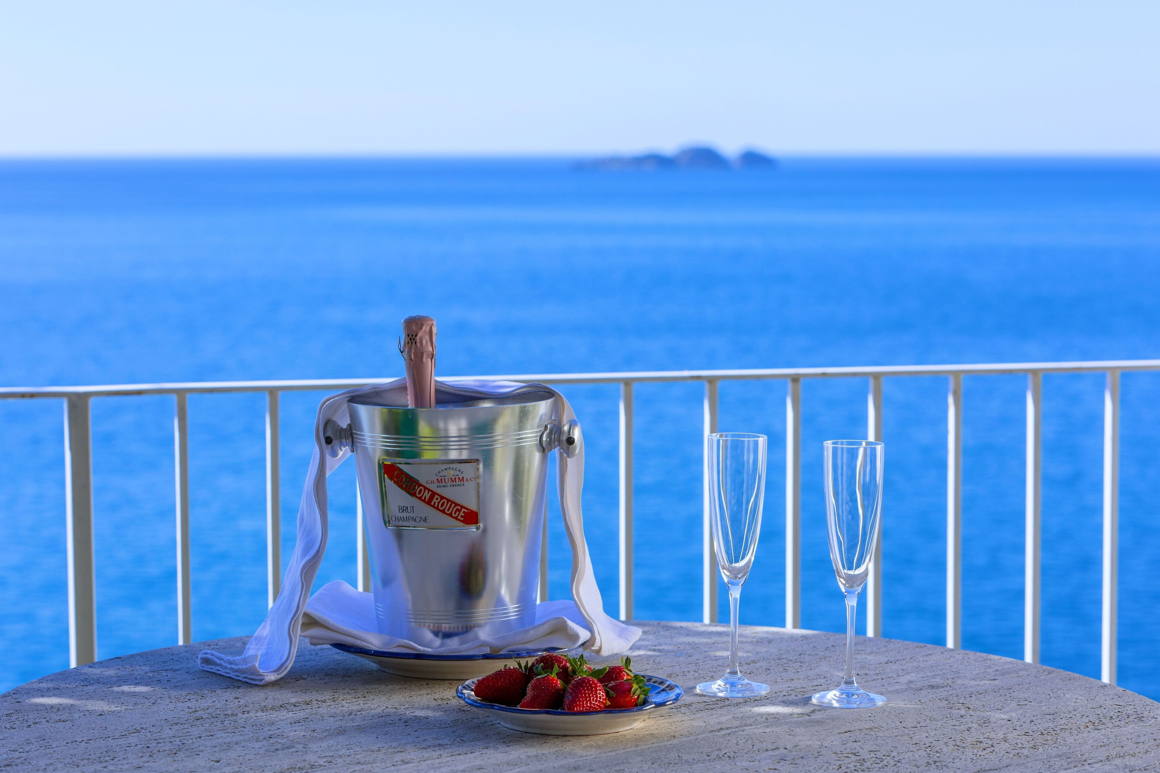 A table with a silver container, two glasses, and a plate of strawberries by a railing with a view of the sea.