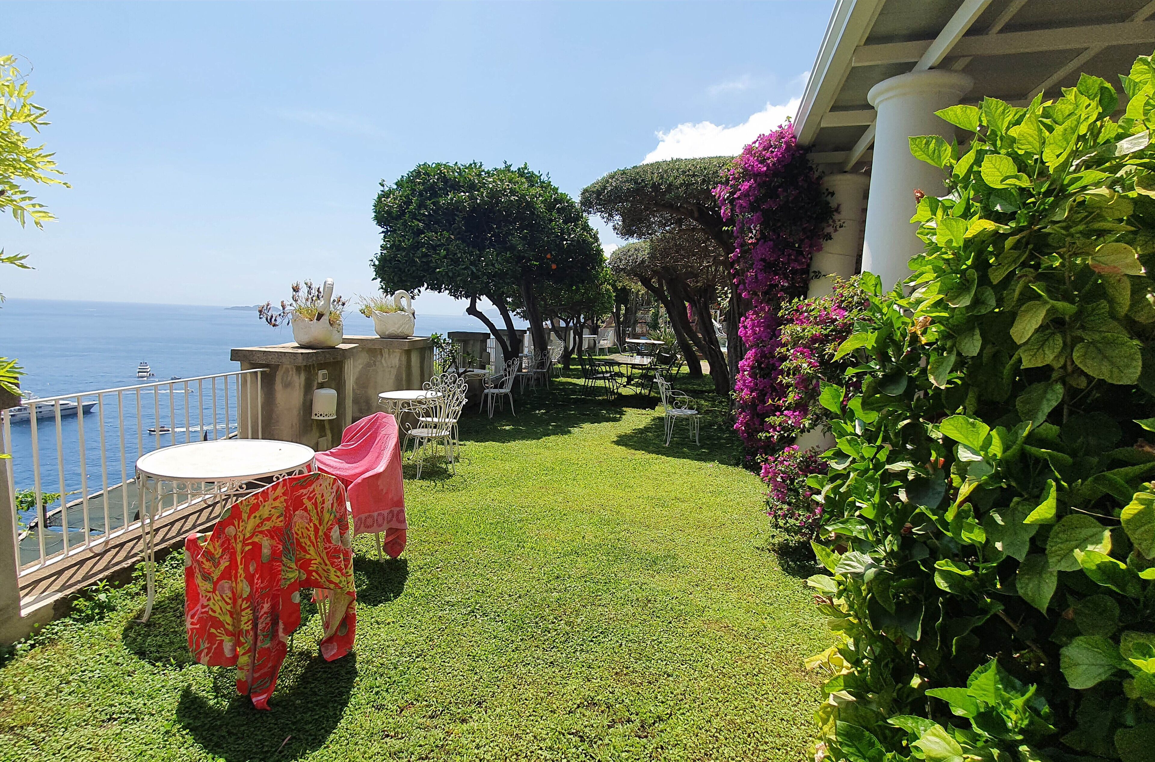 A terrace with a view of the sea, featuring a small table and chairs, surrounded by greenery and a white column.