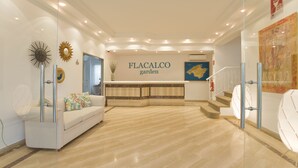 A modern reception area with a white sofa, a glass partition, and a sign reading "FLACALCO garden". - Flacalco Hotel & Apartments (Capdepera)