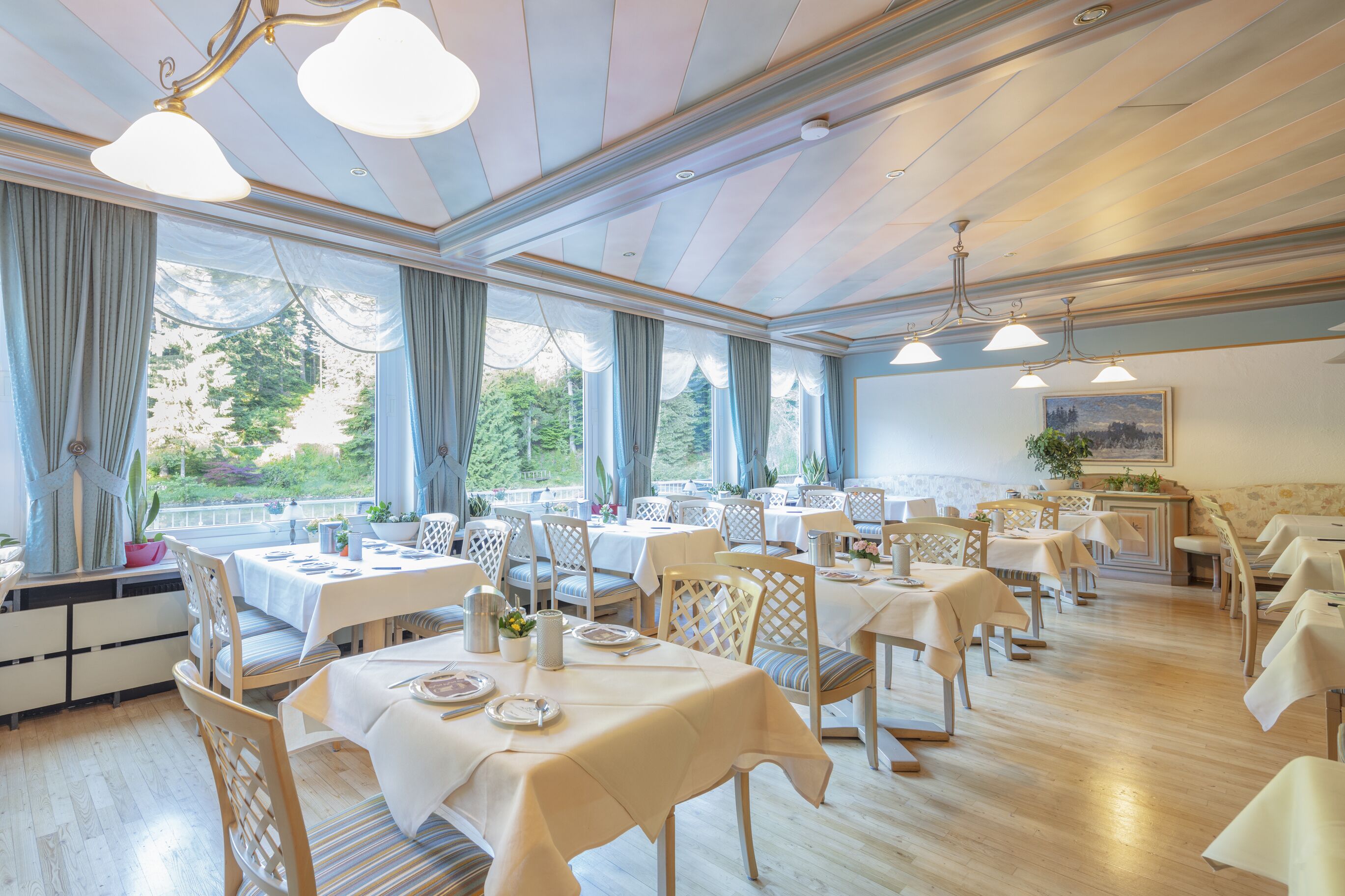 A spacious dining area with multiple tables and chairs, large windows with curtains, and a chandelier.